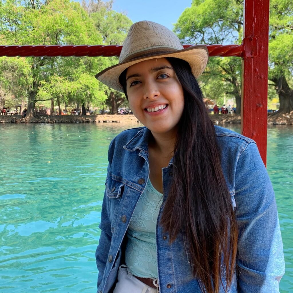 La imagen muestra a perla sentada en una barandilla roja, cerca de un cuerpo de agua. Ella tiene el cabello largo y lacio, y está sonriendo. Lleva puesto un sombrero de paja, una chaqueta de mezclilla azul claro, una blusa de encaje color azul claro, y pantalones claros. El fondo de la imagen es un entorno natural con árboles de hojas verdes y un cielo despejado. El agua detrás de ella es clara y refleja el color del cielo y los árboles, creando un ambiente sereno y relajante. Hay algunas personas en el fondo, disfrutando del área al aire libre.