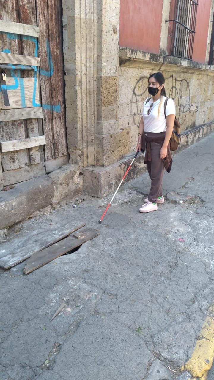 La imagen muestra a perla caminando por una acera en una calle. Ella tiene el cabello largo y oscuro, y lo lleva recogido en una cola de caballo. Viste una camiseta blanca, pantalones marrones, y zapatillas deportivas rosadas. Lleva una chaqueta marrón atada a la cintura y una mochila de tela marrón en la espalda. También usa una mascarilla negra que le cubre la nariz y la boca.En su mano derecha, perla sostiene un bastón blanco con la punta roja, que utiliza para caminar y detectar obstáculos debido a su discapacidad visual. La acera por la que camina está en mal estado, con grietas y un agujero cubierto parcialmente por tablas de madera, lo que representa un peligro potencial.El fondo de la imagen muestra una pared de piedra beige con grafitis y una puerta de madera desgastada con algunas tablas clavadas de forma desordenada. La puerta y la pared tienen un aspecto antiguo y descuidado. A la derecha de la imagen, se ve una parte de la calle con una línea amarilla pintada en el borde de la acera. La escena parece estar en una zona urbana antigua y descuidada.